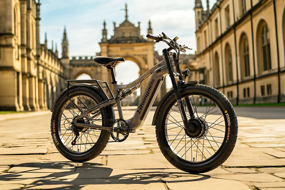 Warm golden-hour static product shot of matte grey Shengmilo S600 full-suspension fat tire e-bike parked on stone pavement before classic European historic architecture.