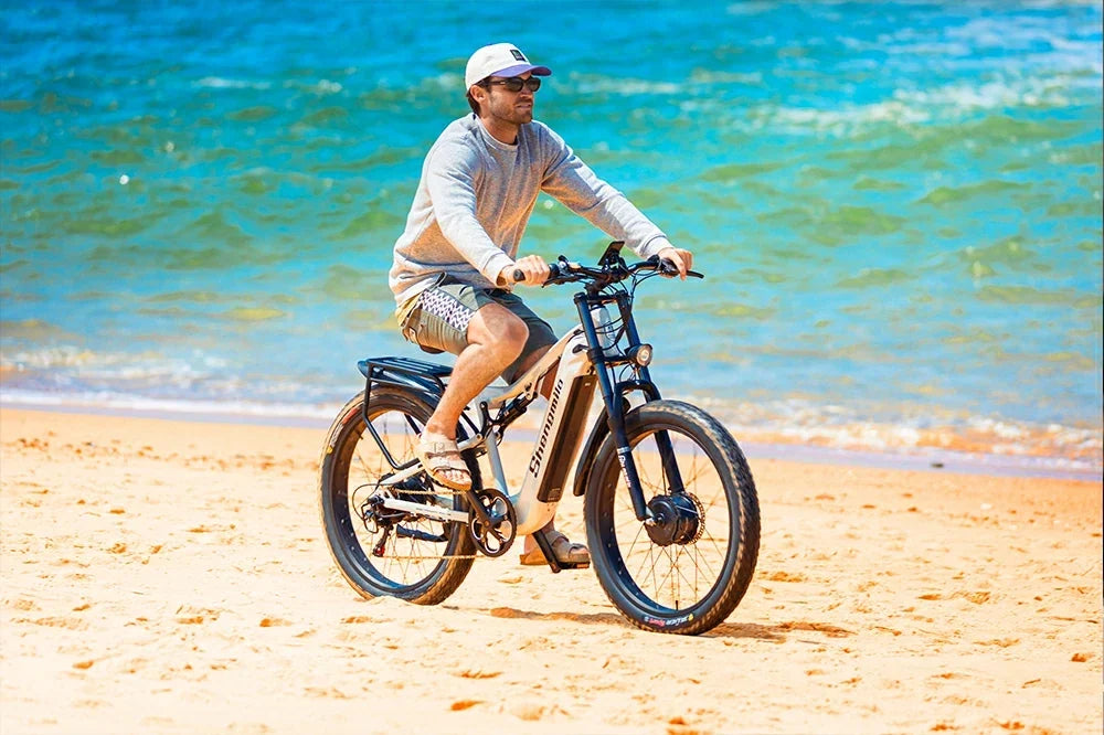 Man riding white Shengmilo S600 full suspension fat tire electric bike along a sunny sandy beach beside turquoise ocean.