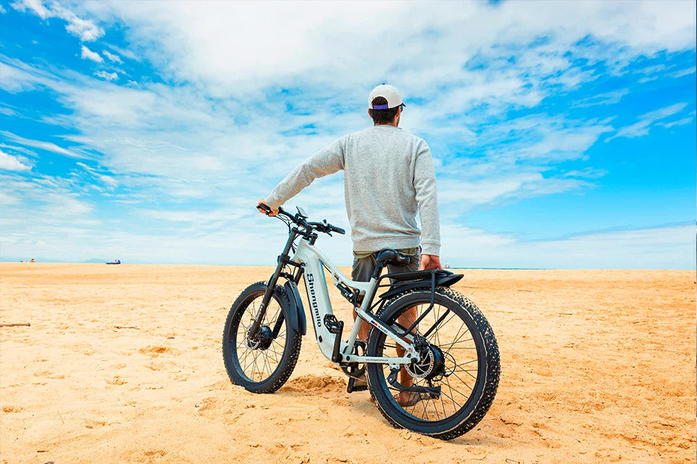 Bright seaside lifestyle shot of a man standing beside light grey Shengmilo S600 fat tire electric bike parked on wide golden sand beach under vivid blue cloud-dappled sky.