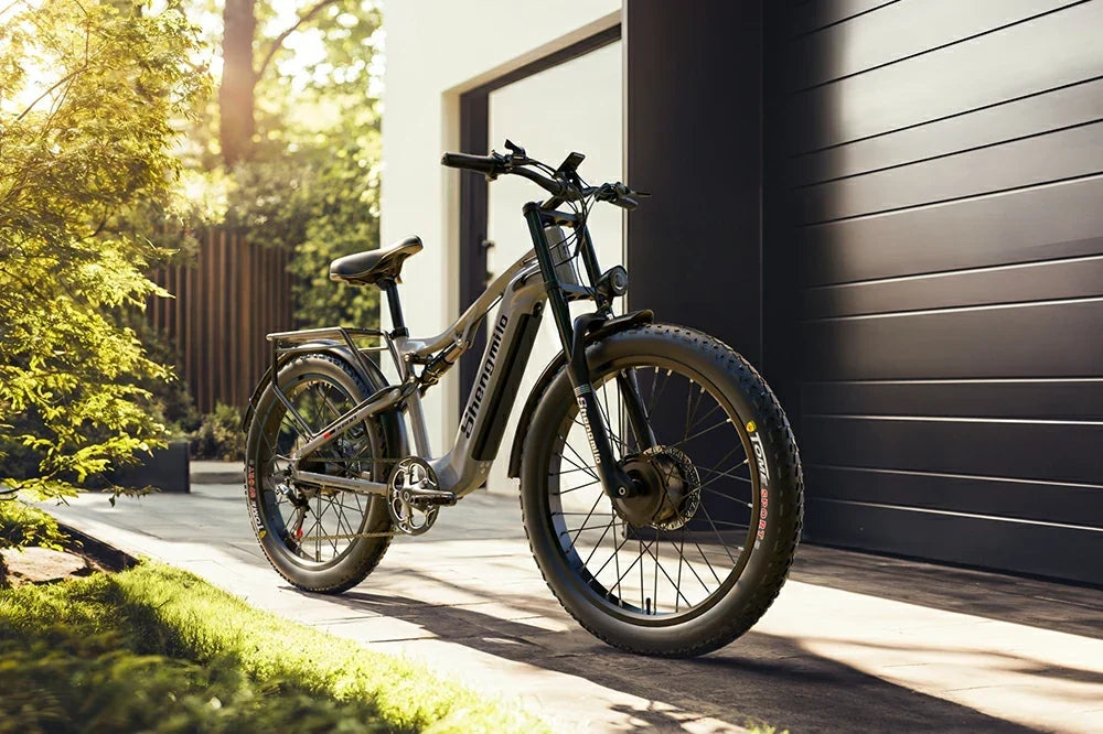 Warm golden-hour static product shot of matte grey Shengmilo S600 full-suspension fat tire e-bike parked on the paved patio outside a modern minimalist home with lush green garden foliage.