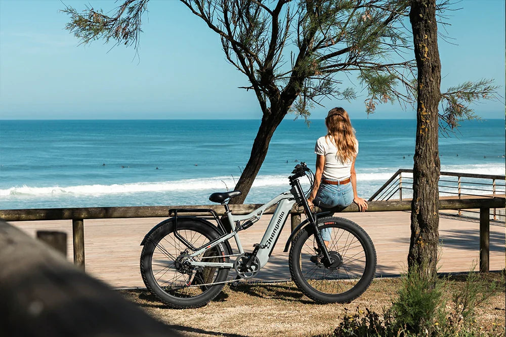 Bright coastal lifestyle shot of a woman leaning on a wooden beach railing beside pale grey Shengmilo S600 fat tire e-bike overlooking turquoise ocean waves.