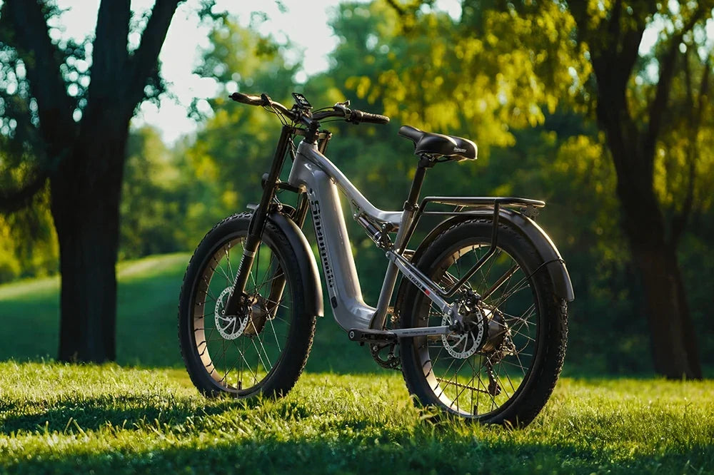Golden hour park static product shot of silver full suspension Shengmilo S600 fat tire electric bike resting on lush green grass amid sunlit leafy trees.