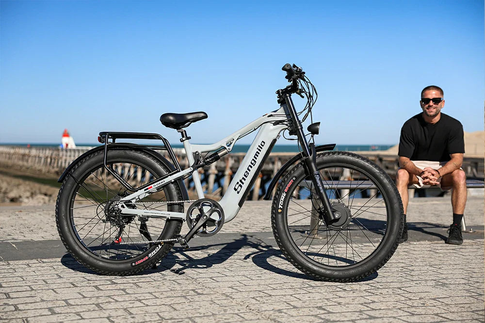 Bright seaside static product shot of light grey Shengmilo S700 full-suspension fat tire e-bike on coastal paved promenade with a seated male model and ocean pier backdrop.
