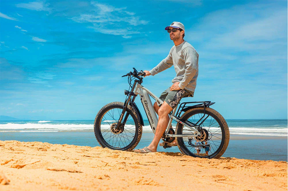 Light grey Shengmilo S600 full suspension all-terrain electric bicycle, male rider beside ebike on golden beach with turquoise ocean and bright blue sky backdrop for coastal outdoor cycling