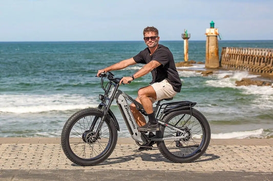 Coastal lifestyle shot of a man posing on light grey Shengmilo S600 fat tire electric bike on seaside stone pavement with ocean and lighthouse backdrop.