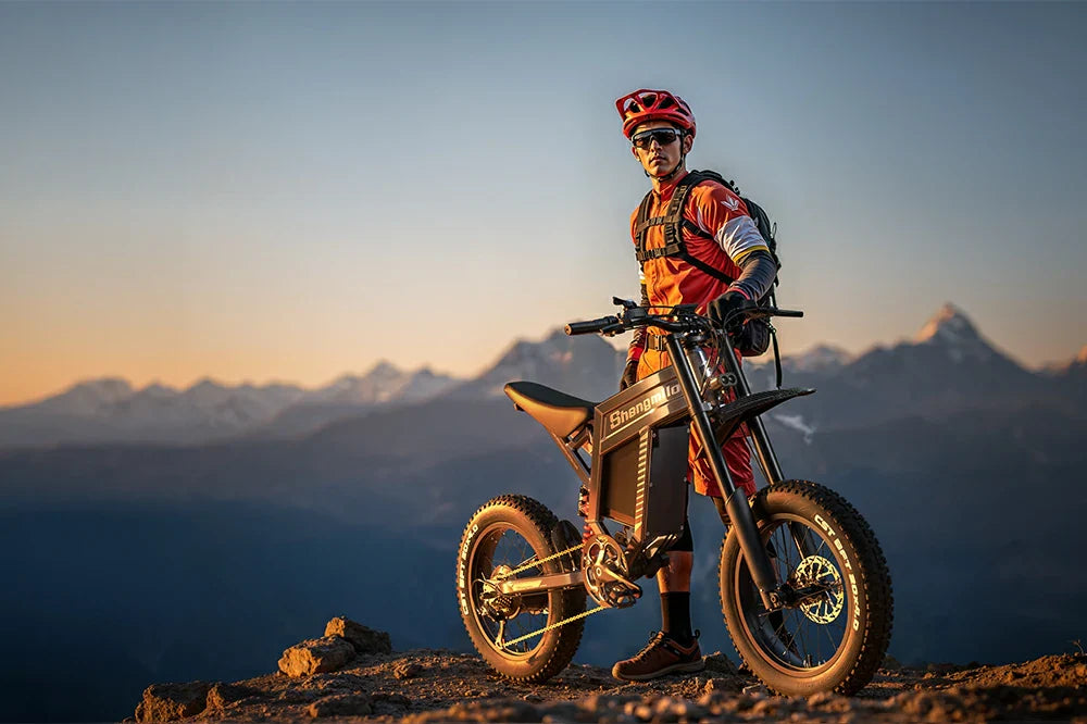 Shengmilo S900 high-performance off-road e-bike with a rider standing atop a mountain at sunset.