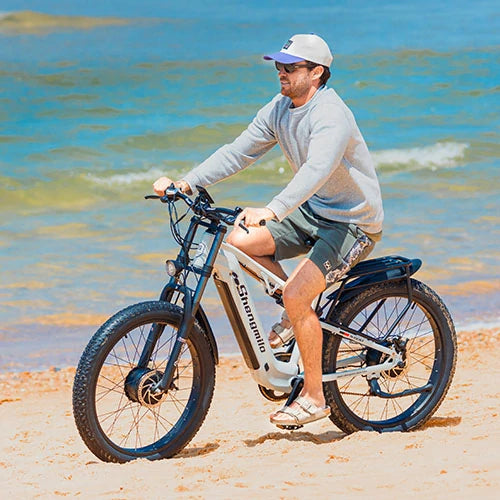 Shengmilo MX06 white full suspension fat tire e-bike, male rider cycling along sunny coastal beach beside blue seawater, all-terrain beach outdoor electric bicycle