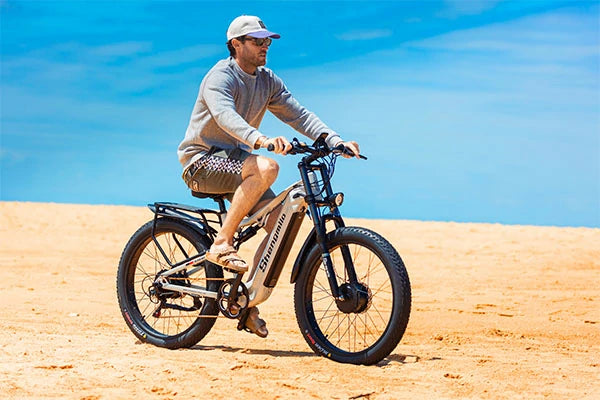 Silver Shengmilo S600 full suspension all-terrain fat tire electric bike, male rider in cap cycling across dry sandy desert under clear bright blue sky for outdoor adventure promotion