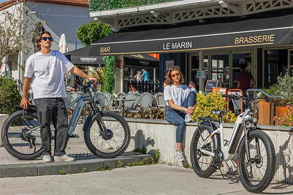 Two color options of Shengmilo S600 and S700 full suspension fat tire e-bikes, man and woman relaxing beside ebikes at outdoor French brasserie sidewalk, European town leisure cycling lifestyle photo