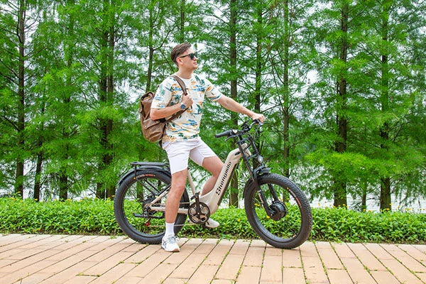 Cream Shengmilo S700 full suspension fat tire electric bicycle, male rider with brown backpack standing on wooden trail in lush green forest park, casual outdoor leisure cycling lifestyle shot