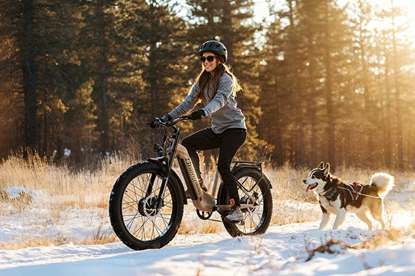 Khaki Shengmilo S700 full suspension all-terrain fat tire electric bike, helmeted female rider cycling on snow-covered forest path, husky dog running alongside in warm sunset winter woods