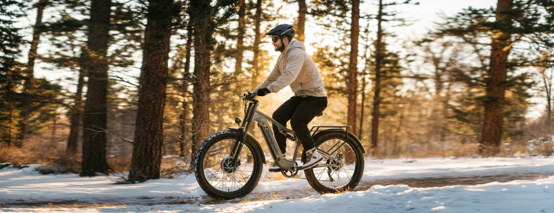 This is the Shengmilo S600, an olive-green full-suspension fat tire electric bike, ridden by a man with helmet and casual wear along a snow-covered forest trail bathed in warm sunset woodland light.