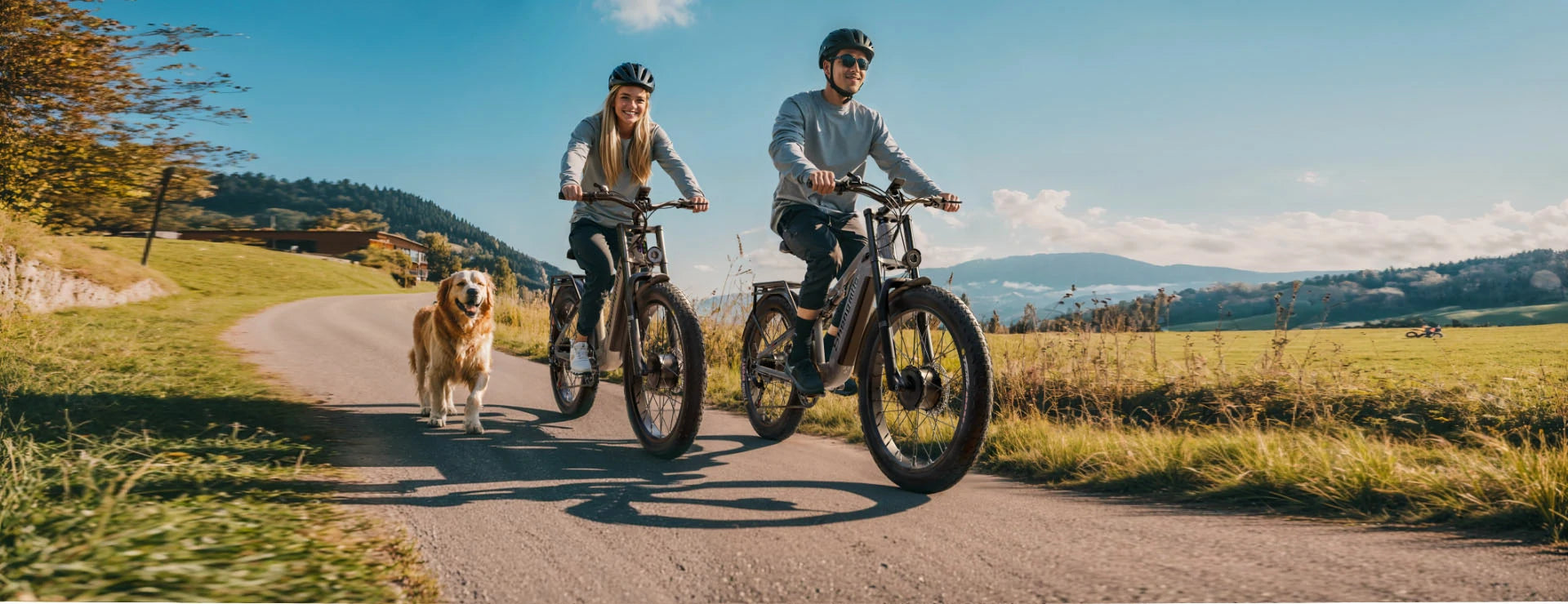 A couple rides Shengmilo S600 & S700 fat-tire electric bikes along a scenic countryside paved trail, with a golden retriever walking beside them. Rolling green meadows, forested hills and clear blue sky form the pastoral backdrop.