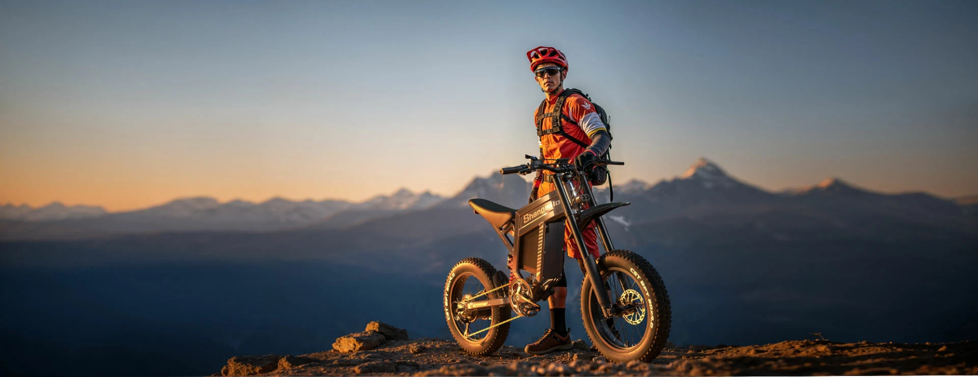 Shengmilo S900 high-performance off-road electric motorcycle style e-bike, rider with red helmet stands on mountain cliff at sunset, snow-capped alpine mountain backdrop for extreme outdoor sports promotion